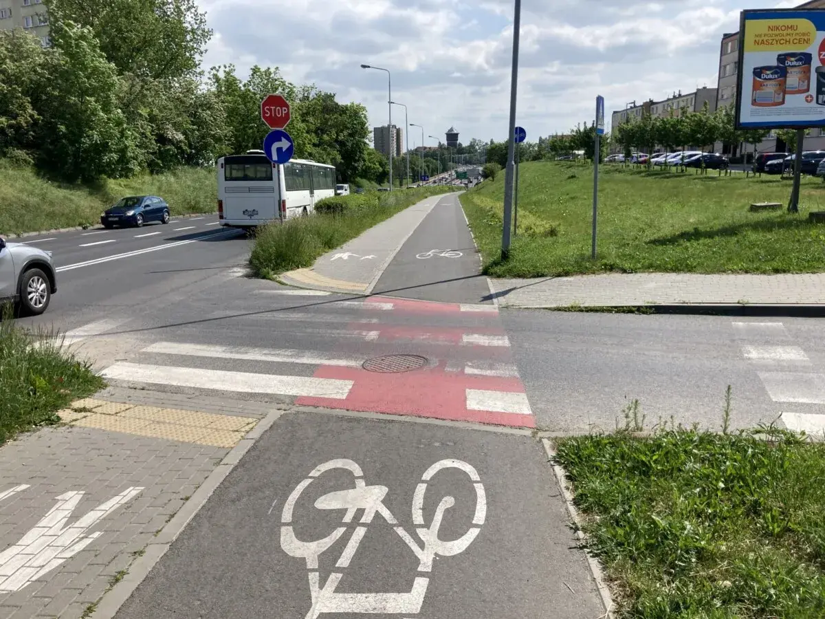 Wyraźna droga dla rowerów z oznakowaniem, obok pasy dla pieszych i jezdnia z autobusem.
