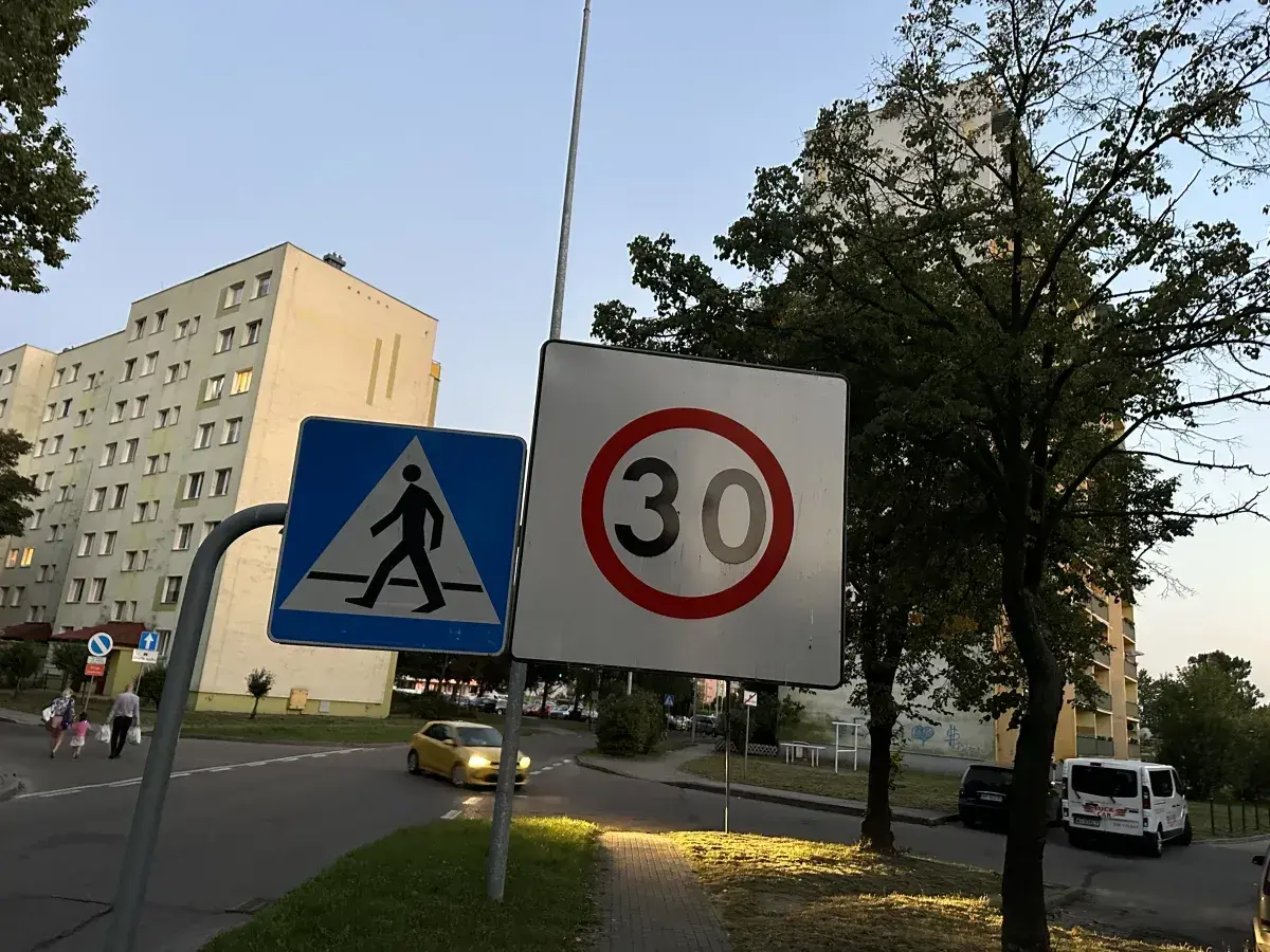 Znak D43 (przejście dla pieszych) i znak ograniczenia prędkości do 30 km/h na tle bloków mieszkalnych i drzew.