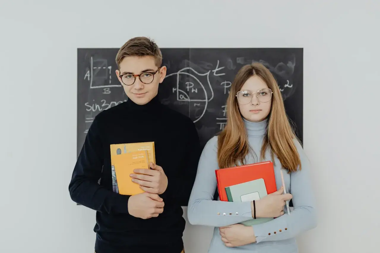 Młodociany uczeń i uczennica z książkami przed tablicą z matematycznymi wzorami.