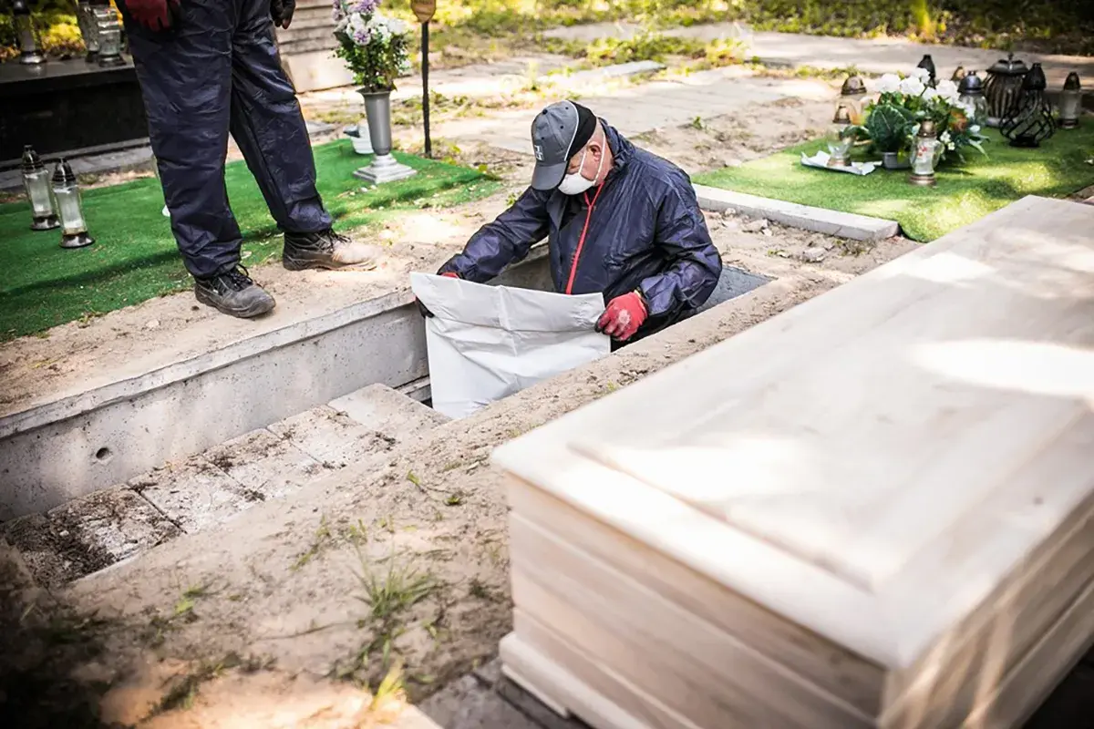 Pracownicy cmentarza przeprowadzają ekshumację, wyciągając biały worek z grobu. Obok leży drewniana trumna.