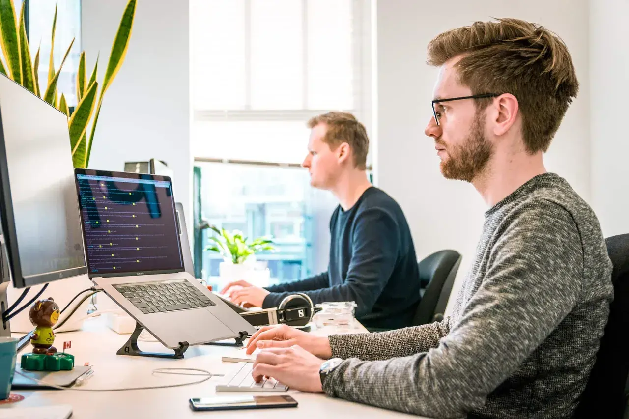 Dwóch programistów pracuje przy komputerach. Jeden z nich ma okulary, co może sugerować refundację okularów przez pracodawcę.