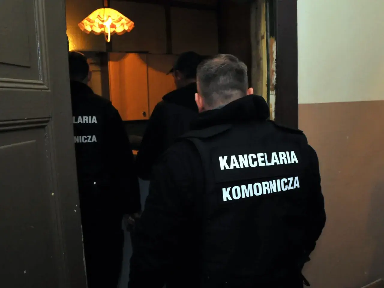 Trzech mężczyzn w czarnych kamizelkach z napisem "KANCELARIA KOMORNICZA" wchodzi do pomieszczenia. Prawdopodobnie to postępowanie egzekucyjne.
