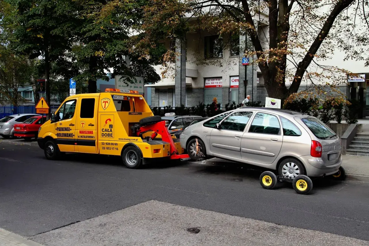 Żółty holownik zabiera srebrny samochód, który zapewne otrzymał mandat za parkowanie.
