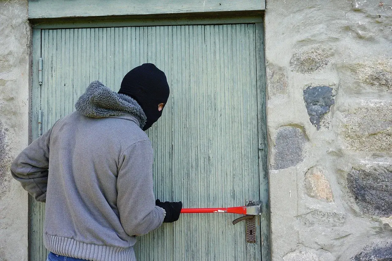 Osoba w masce próbuje wyważyć drzwi łomem. To klasyczny przykład kradzieży z włamaniem.
