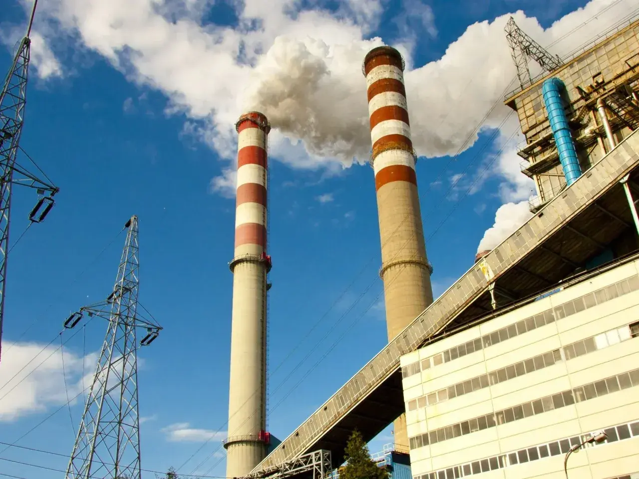 Dwie wysokie, prążkowane kominy elektrowni emitują dym na tle błękitnego nieba. Obok widać linie energetyczne i budynki przemysłowe, tworzące obraz kluczowej infrastruktury.