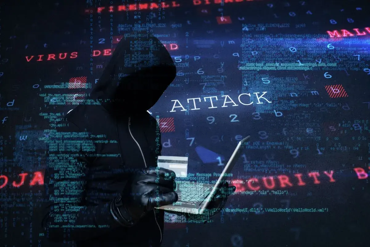 Sylwetka hakera w kapturze, trzymająca laptopa i kartę kredytową. Na tle widać kod i napisy "ATTACK", "VIRUS", "MALWARE", "SECURITY", sugerujące atak hakerski.