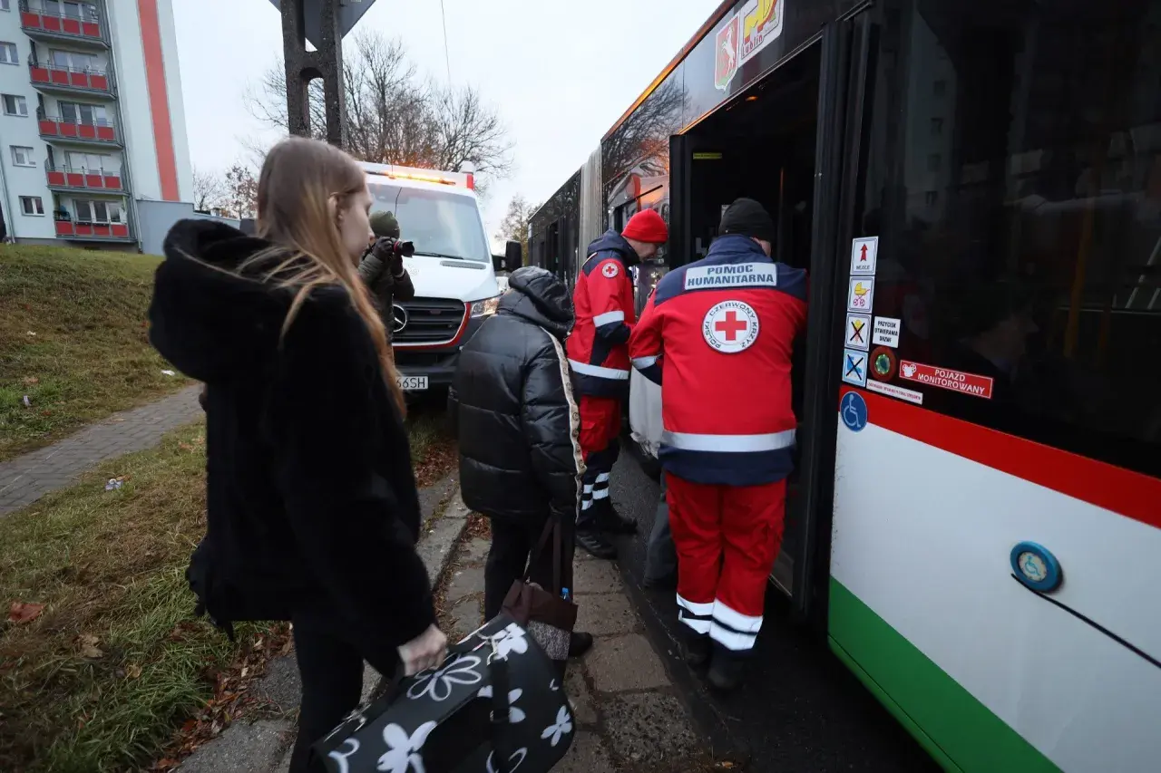 Osoby z bagażami wsiadają do autobusu z pomocą humanitarną. W tle biała furgonetka, przypominająca pojazd z lotniska, gdzie mógł wystąpić alarm.