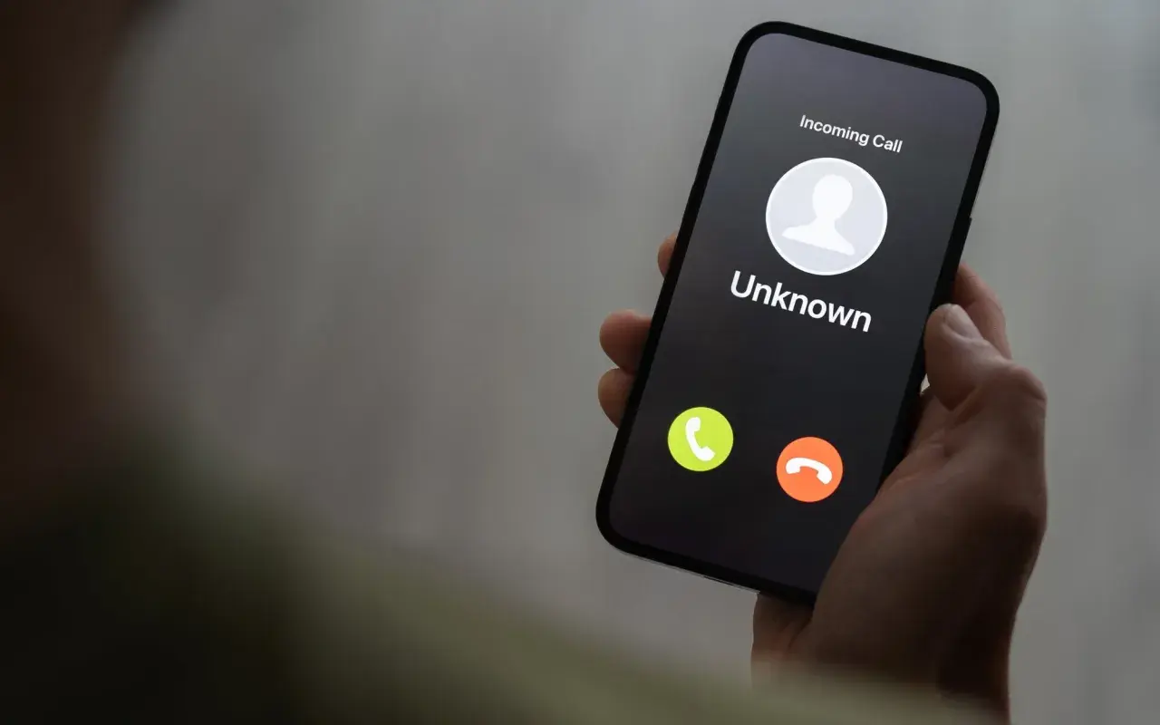 Dłoń trzyma smartfon z ekranem blokady, na którym widnieje "Incoming Call" od nieznanego numeru. Uważaj na vishing!