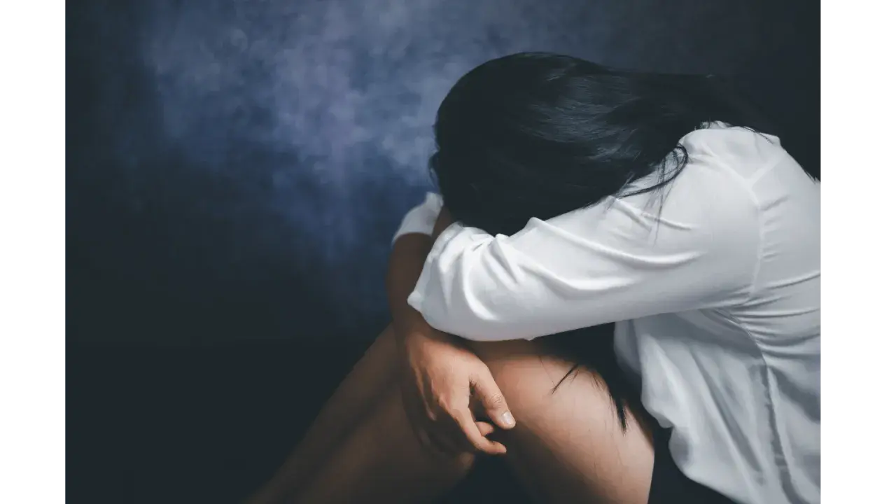 Kobieta w białej koszuli skulona, przeżywająca traumę po gwałcie. Ciemne tło podkreśla jej cierpienie.
