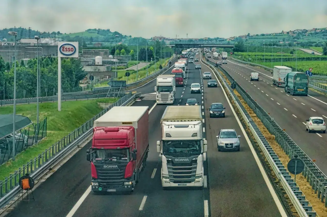 Ciężarówki na autostradzie, jedne wymijają, inne omijają. W tle znak Esso i zielone wzgórza.
