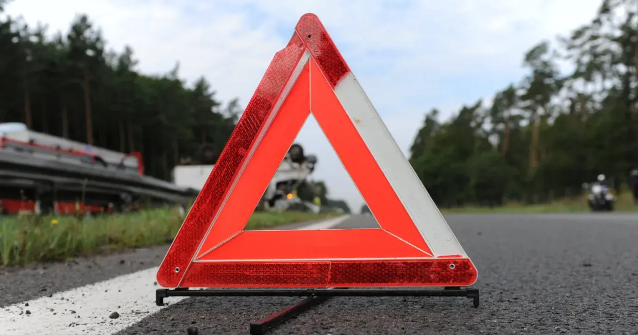 Trójkąt ostrzegawczy, część obowiązkowego wyposażenia samochodu, stoi na poboczu drogi. W tle widać przewrócony samochód ciężarowy.