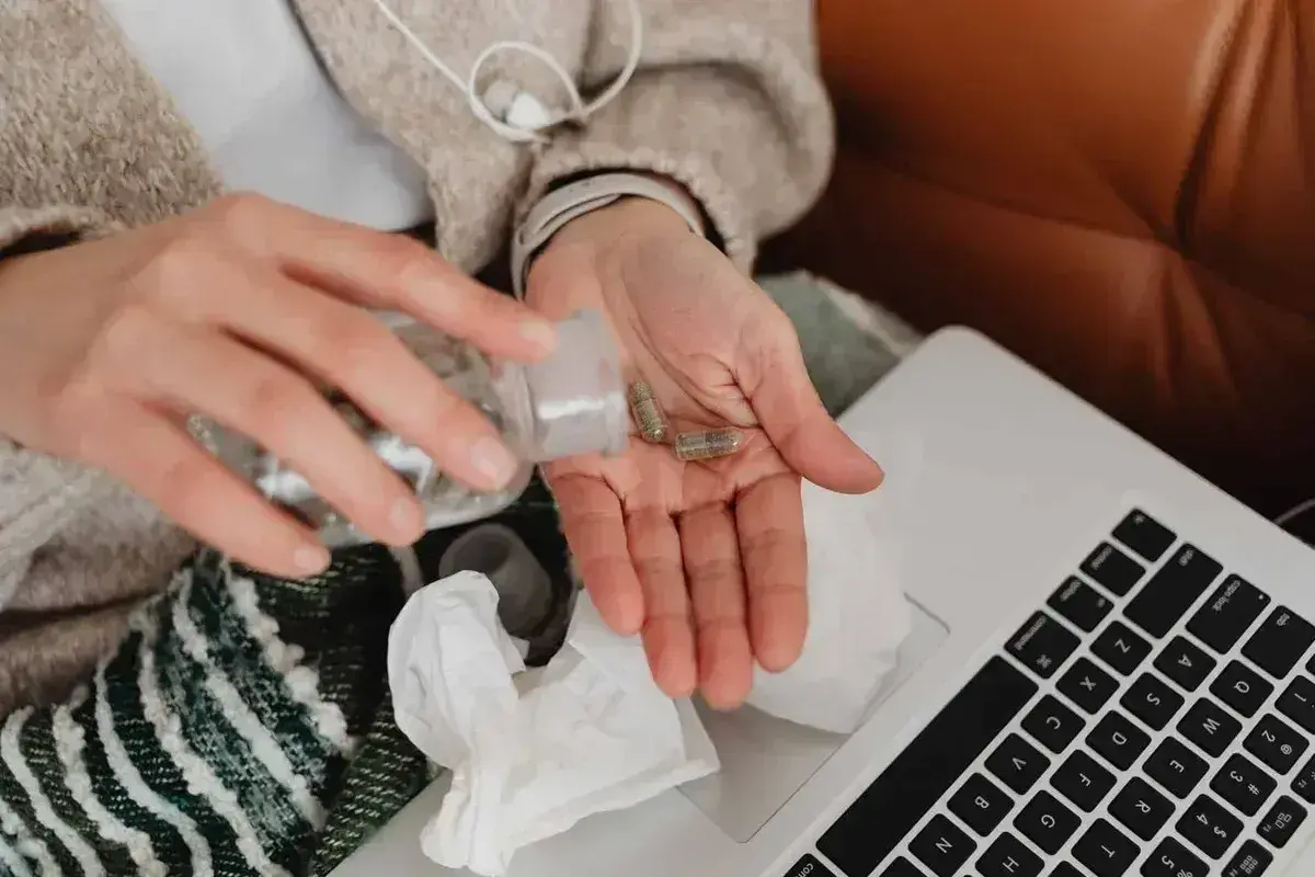 Dłonie z kapsułkami leków, obok laptop i chusteczki. Kto płaci za L4, gdy choroba dopada w domu?