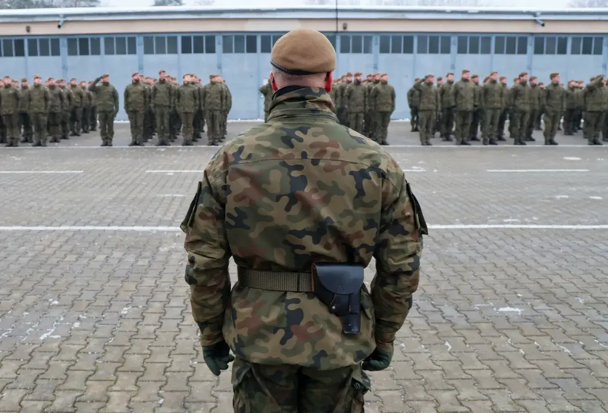 Żołnierz w mundurze moro i beretcie stoi na placu apelowym przed szeregiem innych żołnierzy.