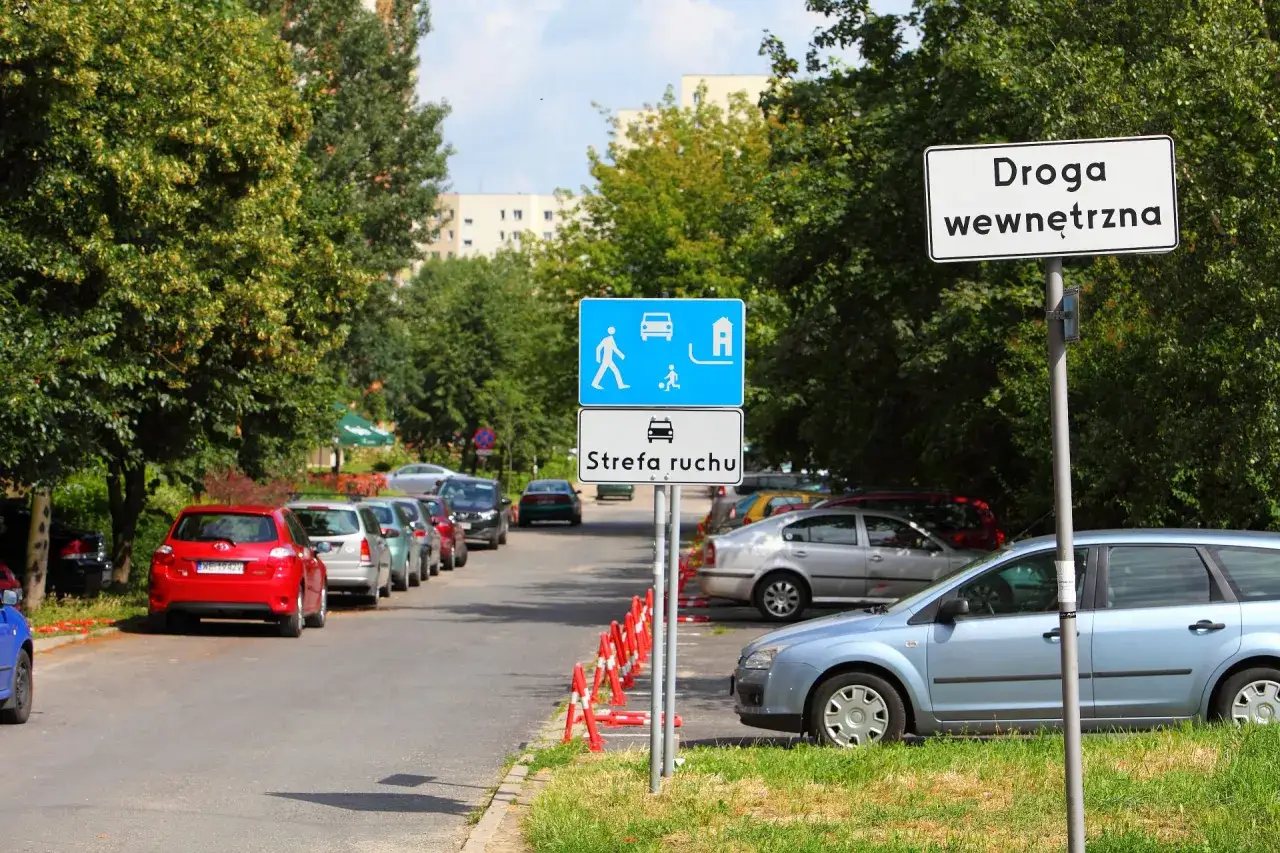 Droga wewnętrzna z oznaczeniem strefa zamieszkania. Samochody parkują wzdłuż ulicy, wśród zieleni.