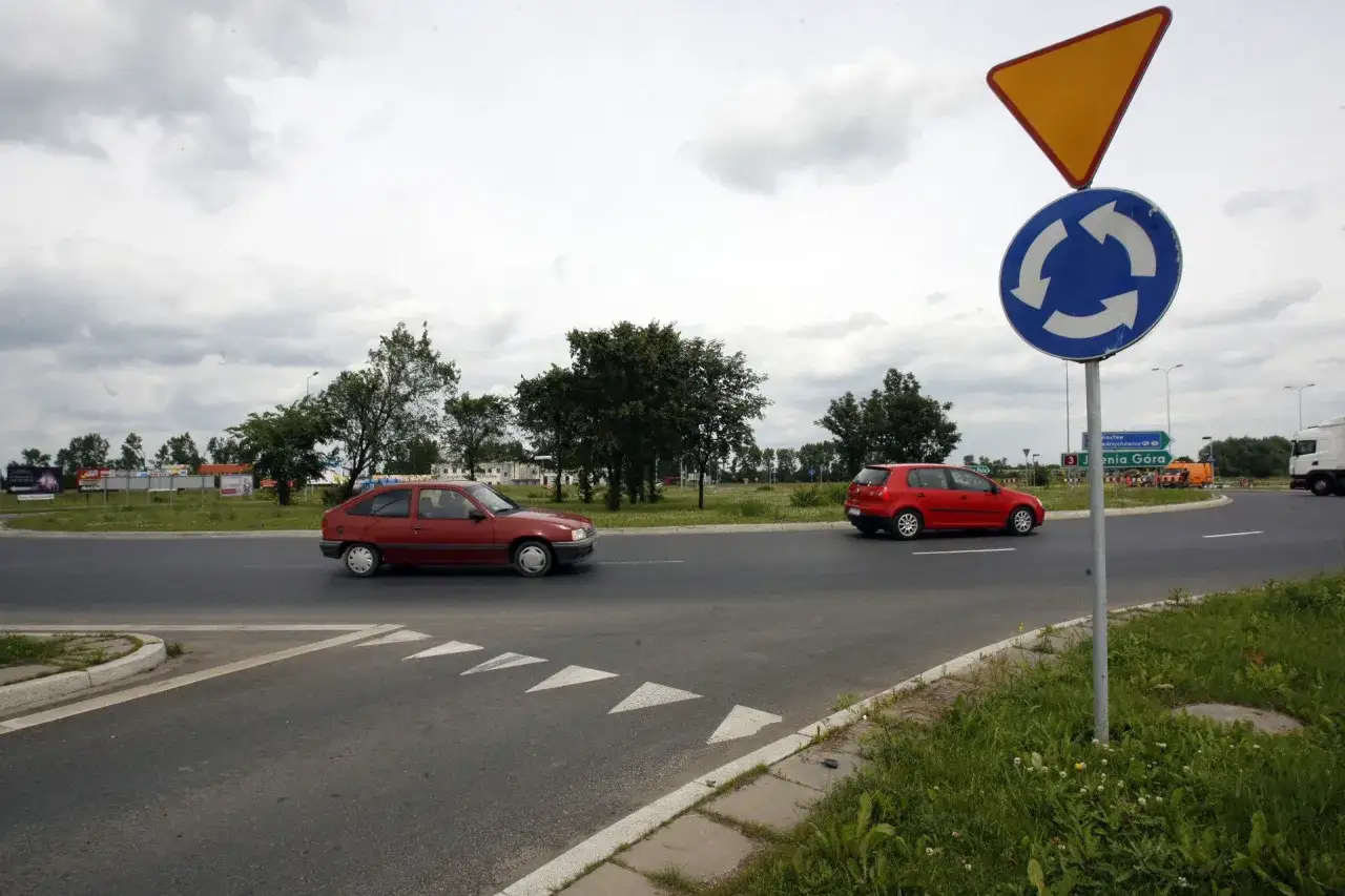 Znak ronda i żółty trójkąt informują, kto ma pierwszeństwo na rondzie. Dwa czerwone samochody zbliżają się do skrzyżowania.