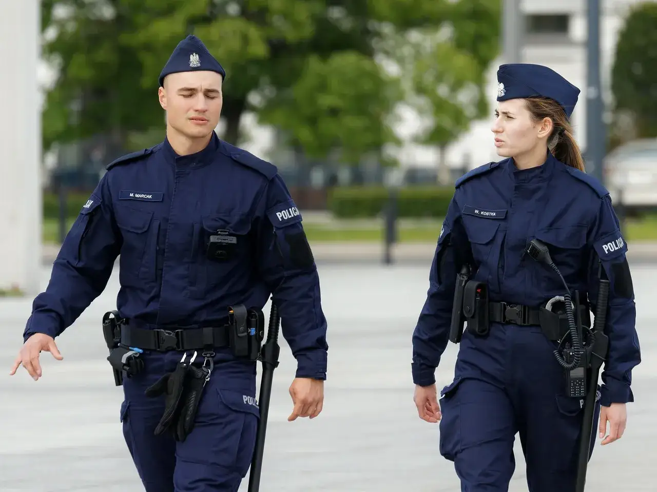 Dwóch policjantów w mundurach idzie ulicą. Mężczyzna, przełożony, ma na pasie rękawiczki i pałkę.
