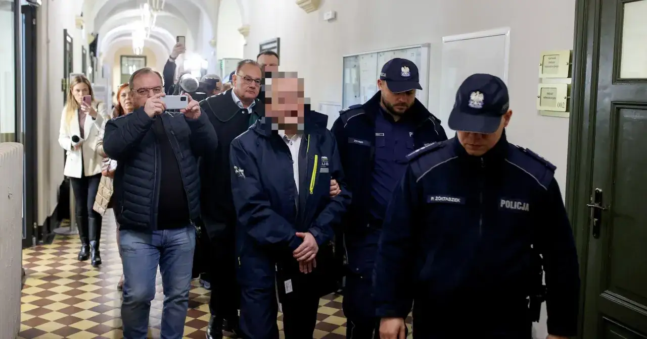 Osoba w niebieskiej kurtce, której twarz jest zamazana, prowadzona przez policjantów w korytarzu sądowym. W tle widać prawnika i osoby z telefonami, być może dokumentujące sprawę związaną z art. 200 kk.
