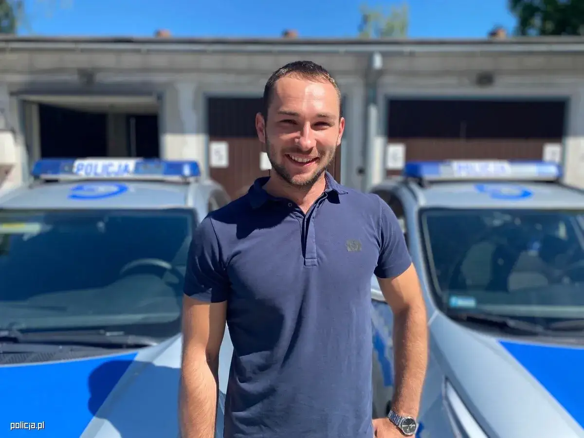 Uśmiechnięty mężczyzna w granatowej koszulce polo stoi przed radiowozem. Czy zastanawiałeś się, ile zarabia pracownik cywilny w policji?