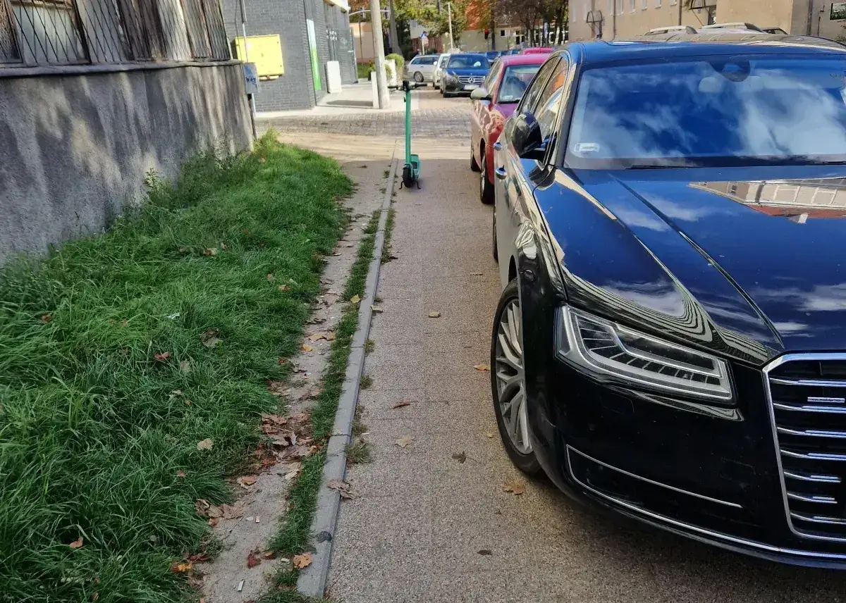 Czarny sedan i czerwony samochód blokują chodnik, utrudniając przejście. Zielona hulajnoga stoi obok, czekając na swojego użytkownika. Parkowanie na chodniku to problem.