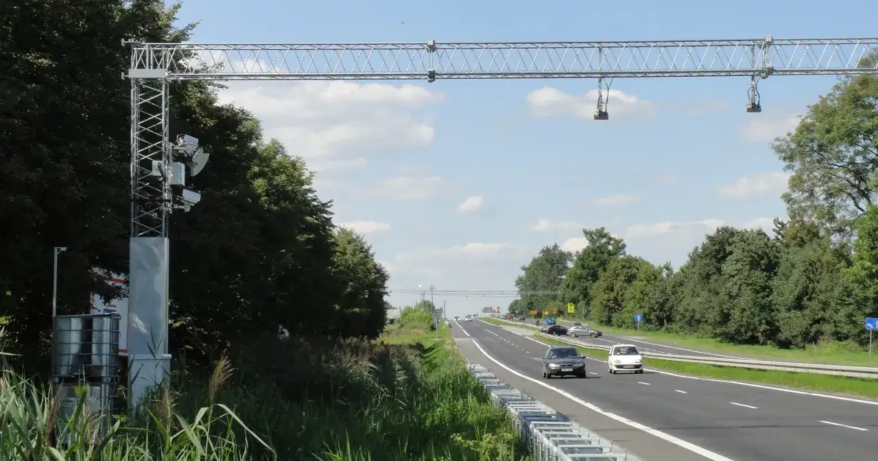 Nad drogą widać konstrukcję do odcinkowego pomiaru prędkości. Samochody jadą po wielopasmowej drodze otoczonej zielenią.