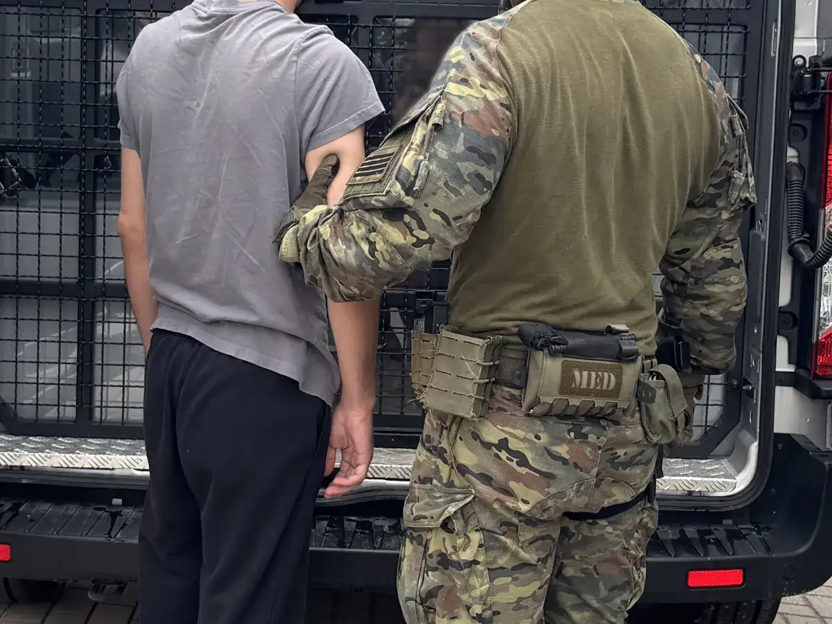 Funkcjonariusz w mundurze taktycznym eskortuje mężczyznę w szarym t-shircie. Sytuacja może być związana z deportacją.