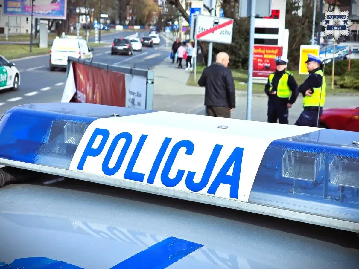 Policyjny kogut z napisem POLICJA. W tle policjanci i pieszy na ulicy.