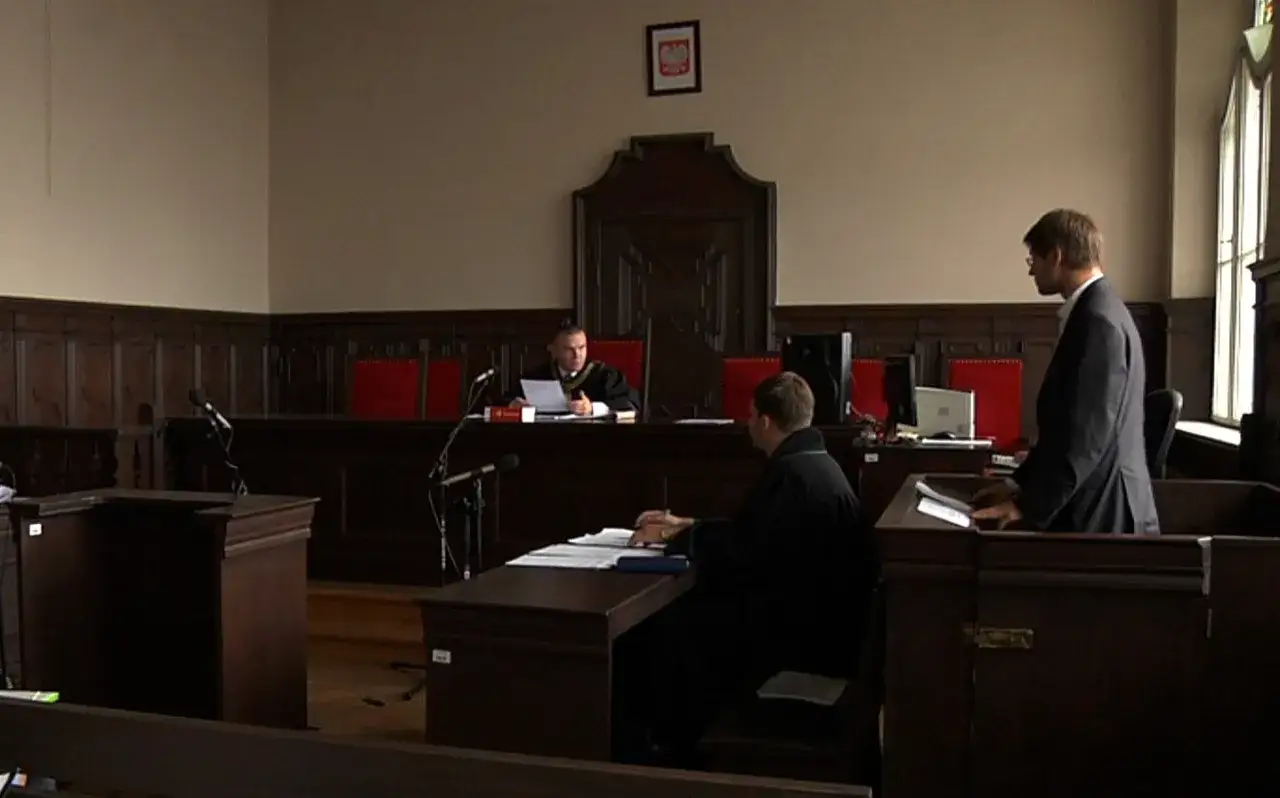 Sądowa rozprawa, sędzia w todze czyta dokumenty, obok siedzi protokolant, a przed nim stoi adwokat. Może to być sprawa z art. 60 kk.