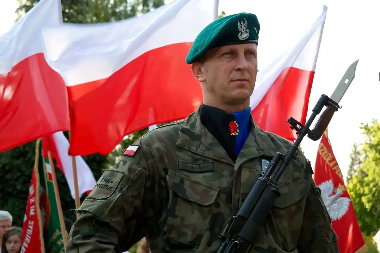 Chorąży w zielonym beretcie, z karabinem i polskimi flagami w tle.