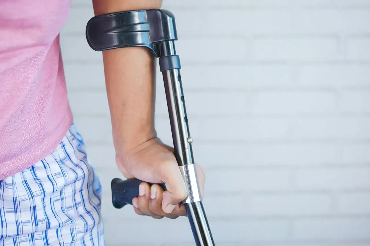Osoba w różowej koszulce i kratkowych spodenkach opiera się na kuli łokciowej. Dłoń pewnie chwyta uchwyt. To może być początek powrotu do zdrowia po urazie, jak np. złamanie nogi, które wymaga wsparcia zgodnie z art. 157 kk.