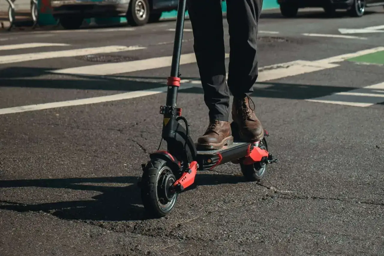 Osoba na hulajnodze elektrycznej na przejściu dla pieszych. Czy hulajnogą elektryczną można jeździć po chodniku? Przepisy są różne.