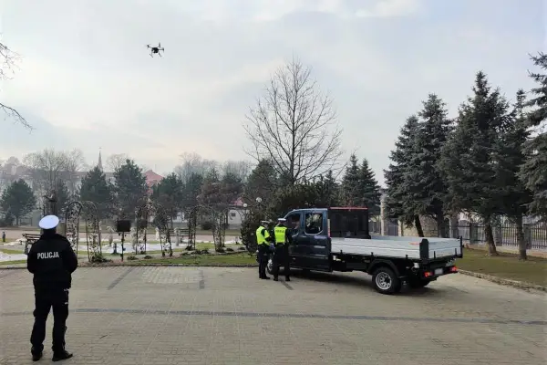 Dron policyjny w powietrzu, obok radiowozu i policjantów.